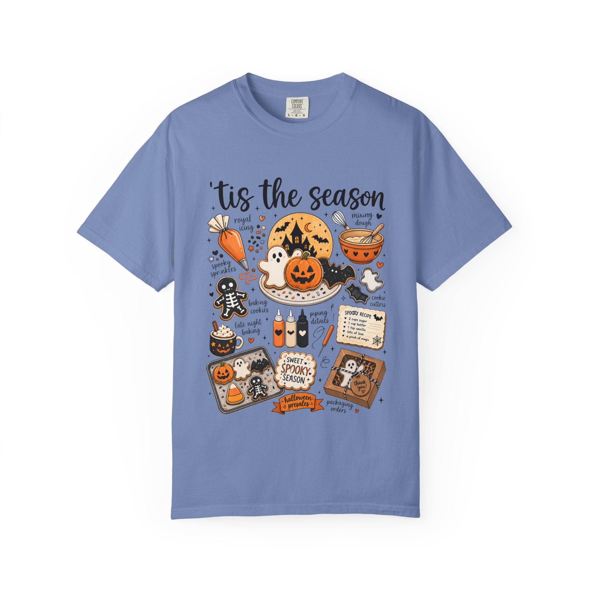 A light blue crewneck t-shirt featuring a colorful Halloween-themed graphic print with the text 'tis the season' and illustrations of jack-o'-lanterns, ghosts, candy canes, and a spider on a white background.