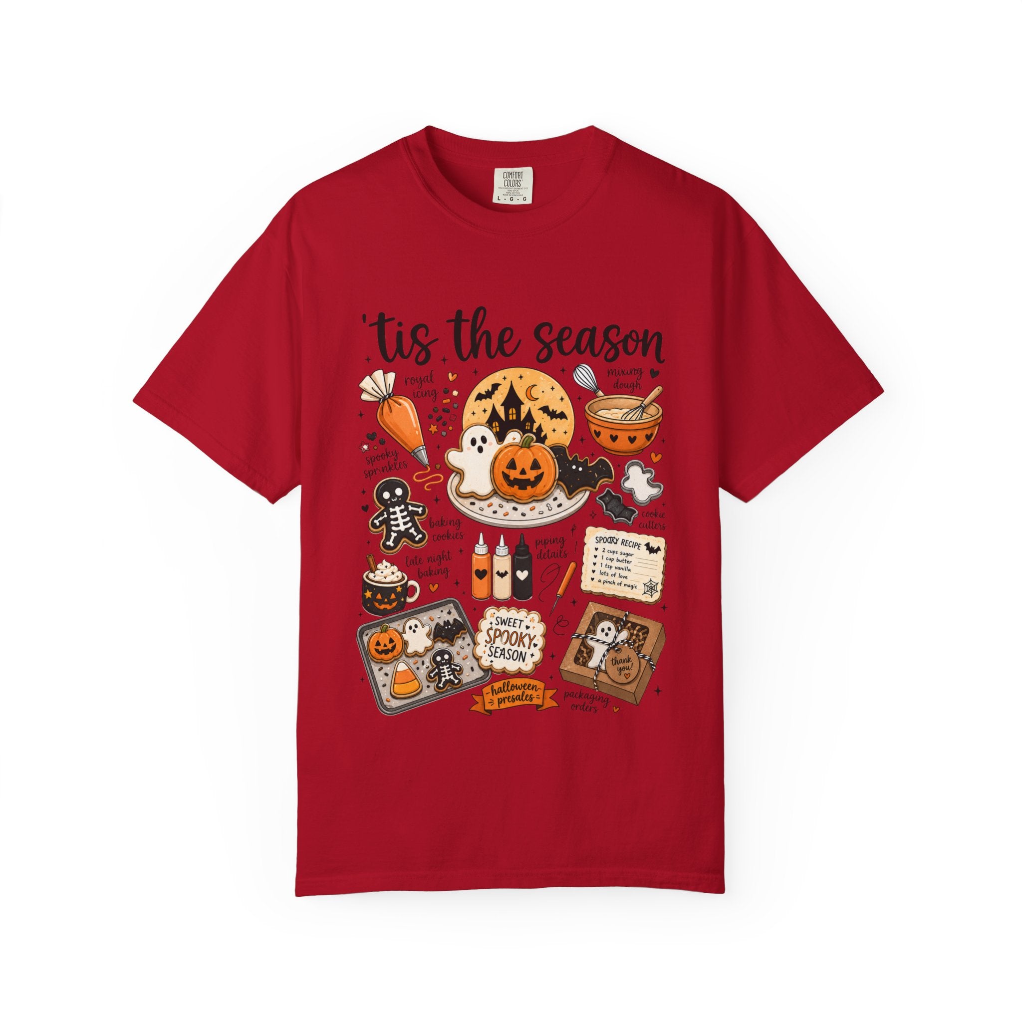 A red short-sleeve crewneck t-shirt featuring a colorful Halloween-themed graphic print on the front with the text 'tis the season' and illustrations of pumpkins, ghosts, treats, and a spiderweb.