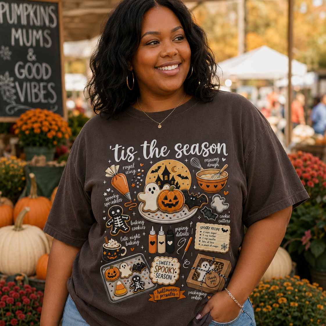 A woman wearing a dark brown graphic t-shirt with a Halloween-themed print that reads 'tis the season' and 'Spooky Spooktacular', paired with light blue jeans, standing in front of an autumn outdoor market background with pumpkins and flowers.