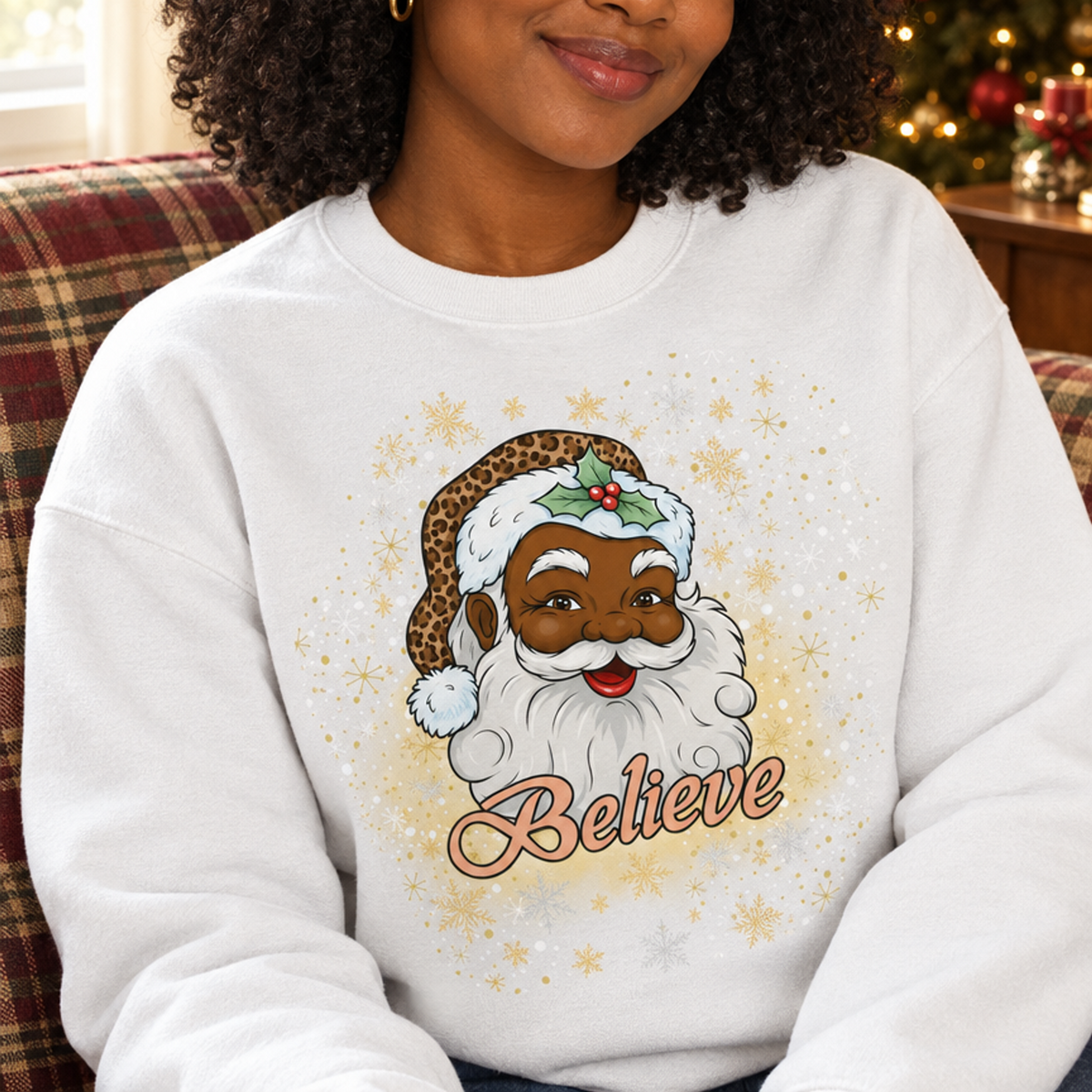 A woman with curly dark hair wearing a white crewneck sweatshirt with a graphic of a Black Santa Claus and the word 'Believe' in script, sitting in a cozy indoor setting with a Christmas tree in the background.