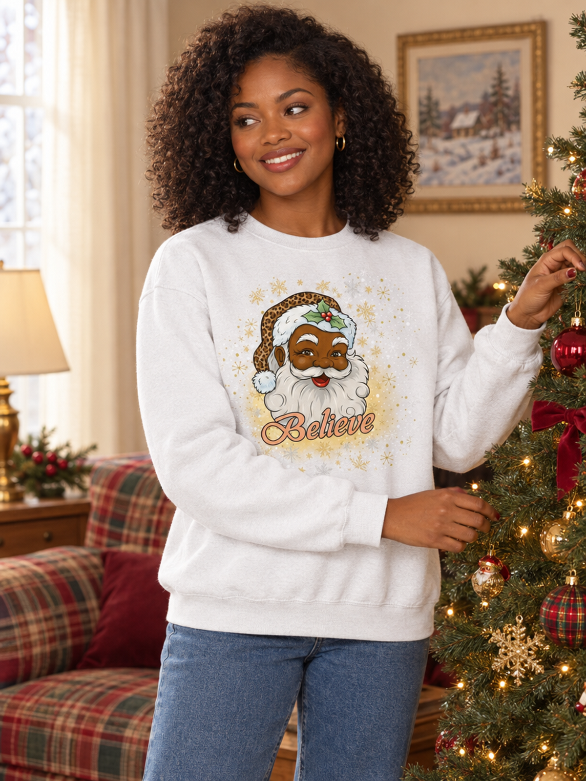A woman wearing a white Christmas sweatshirt with a festive Santa graphic and the word 'Believe' printed on the front, standing in a decorated living room next to a Christmas tree.