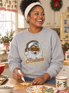 A woman smiling in a cozy kitchen while wearing a light gray crewneck sweatshirt with a festive graphic of Santa Claus and the word 'Believe' in gold glitter-style lettering. She is decorated with a white headband and appears to be icing cookies on a wooden table.