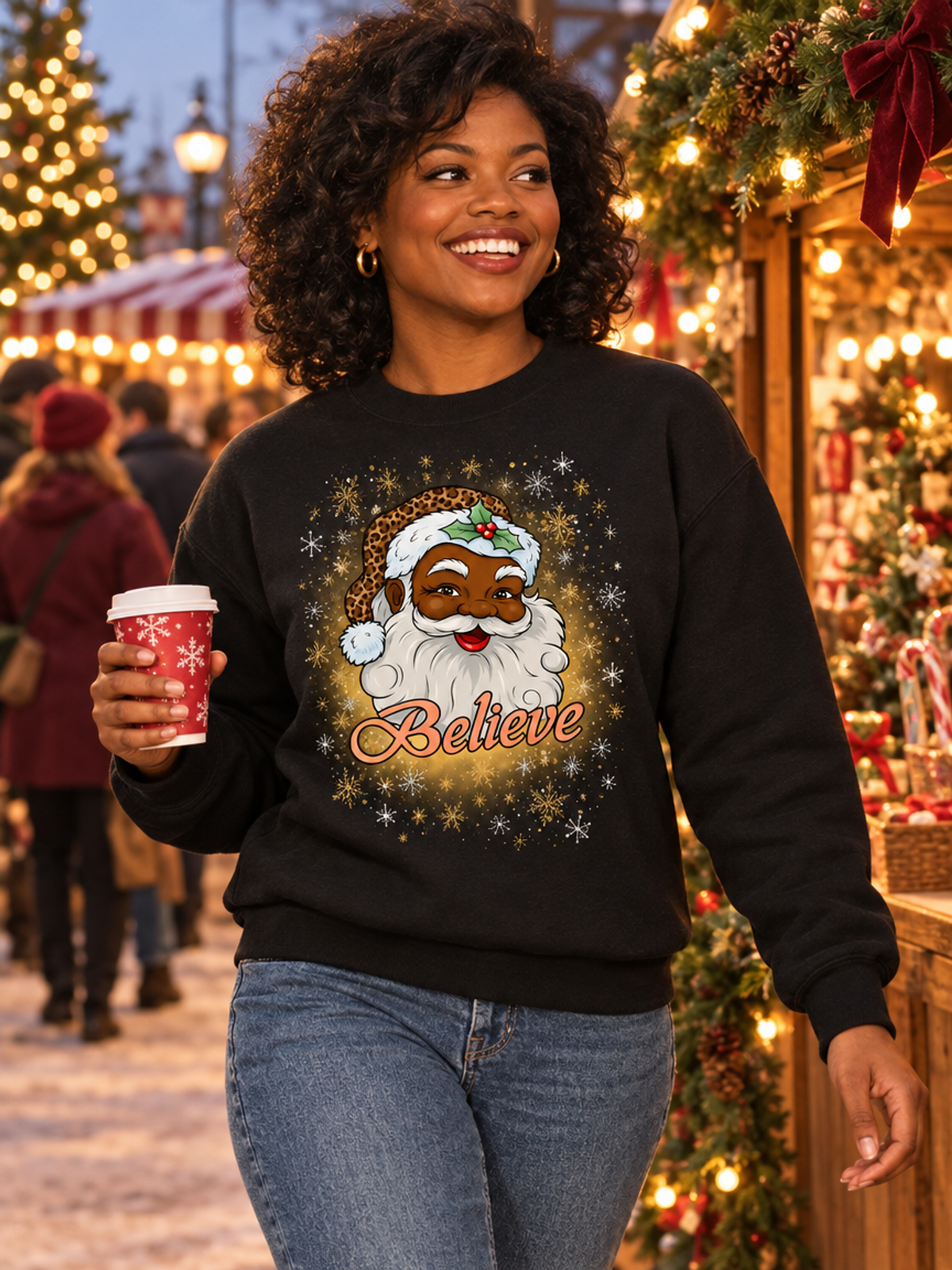 A woman wearing a black crewneck sweatshirt with a festive graphic of a smiling Black Santa Claus surrounded by snowflakes and the word 'Believe' in gold script. She is holding a red Starbucks coffee cup in a decorated Christmas market setting.