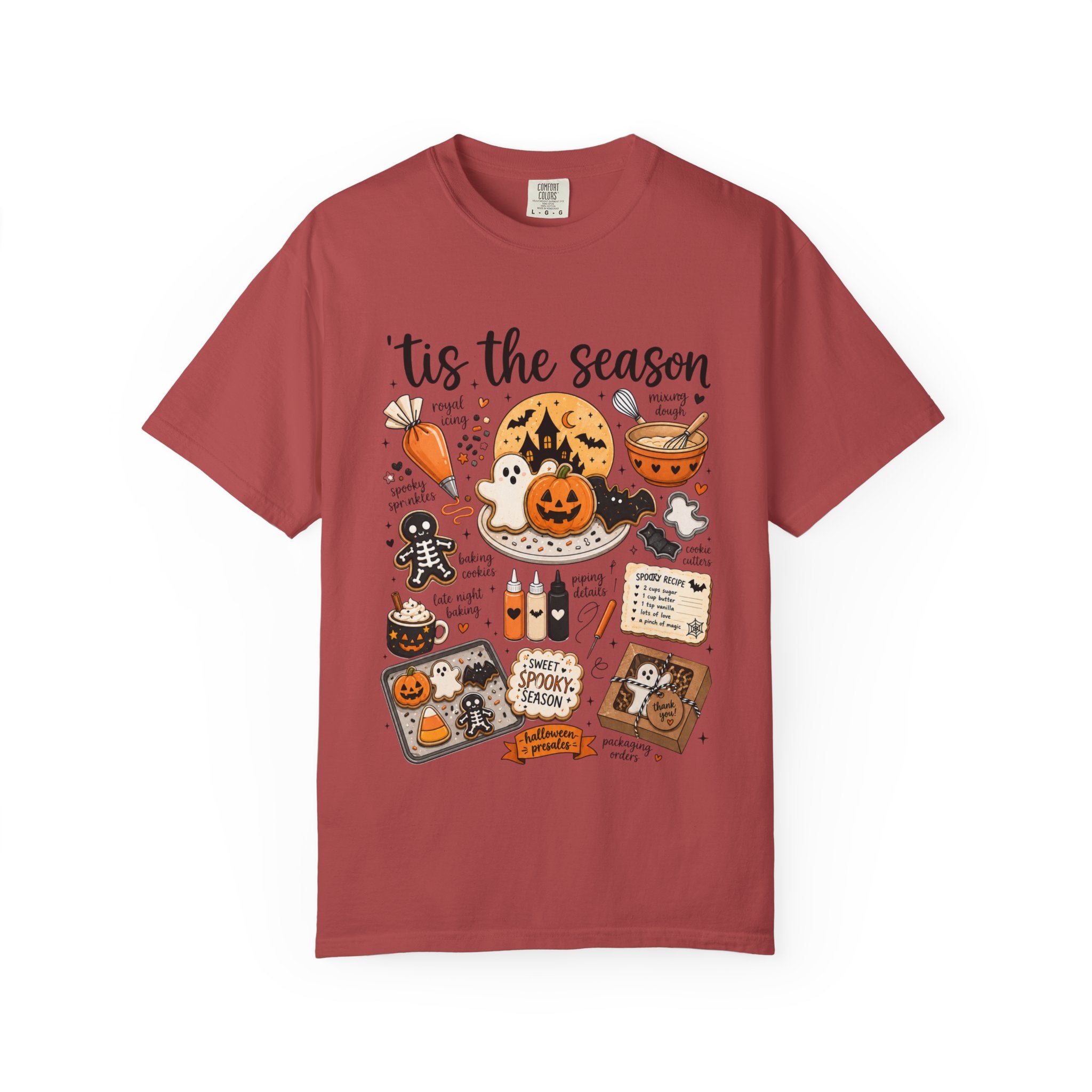 A short-sleeve crewneck t-shirt in a dusty rose or muted brick red color, featuring a detailed Halloween-themed graphic print on the front with the text 'tis the season' and illustrations of jack-o'-lanterns, ghosts, a spider, and a witch's hat.