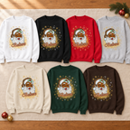 Eight crewneck sweatshirts in different colors are arranged in two rows on a beige fabric surface with a wooden floor and small holiday ornaments visible. Each sweatshirt has a festive Santa face graphic with the word 'Believe' underneath.