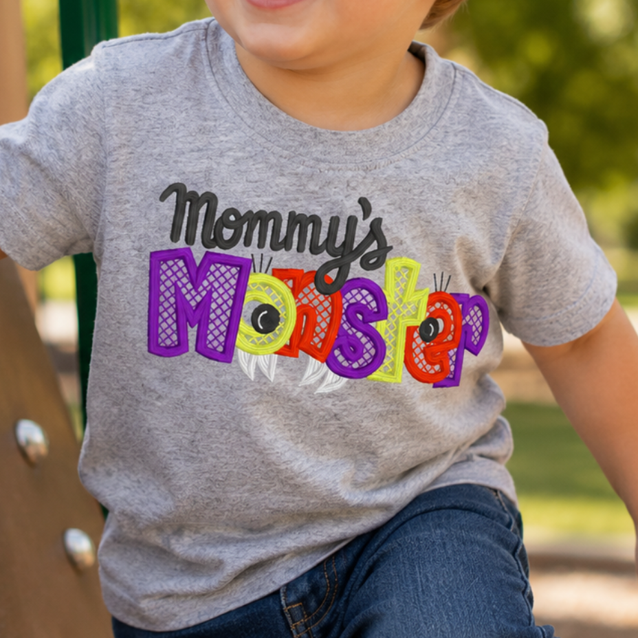 A young boy smiling while climbing a playground slide, wearing a heather gray t-shirt with a colorful 'Mommy's Monster' graphic and blue jeans.