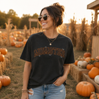 A woman wearing a black graphic t-shirt with 'MOMSTER' printed in orange and white across the chest, standing in a pumpkin patch at sunset.