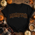 A black short-sleeve T-shirt laid flat on a rustic wooden surface, surrounded by pumpkins, autumn leaves, a plaid scarf, and a mug of coffee. The shirt has the word "MOMSTER" printed across the chest in large orange outlined letters with small spider and bat graphics around it.