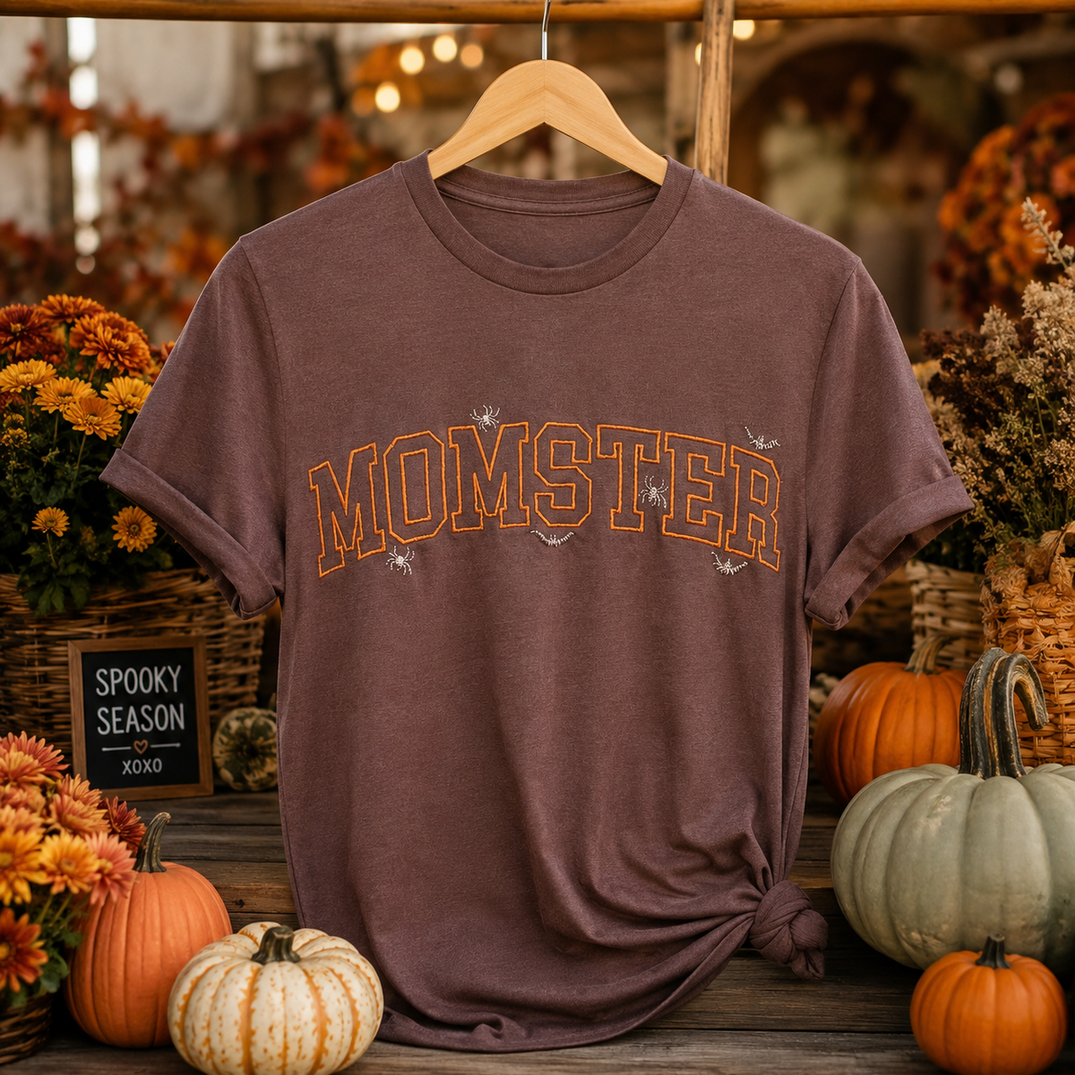 A brown short-sleeve t-shirt with 'MOMSTER' printed in orange arched letters across the chest, hanging on a wooden hanger in a fall-themed setting with pumpkins and autumn decor.