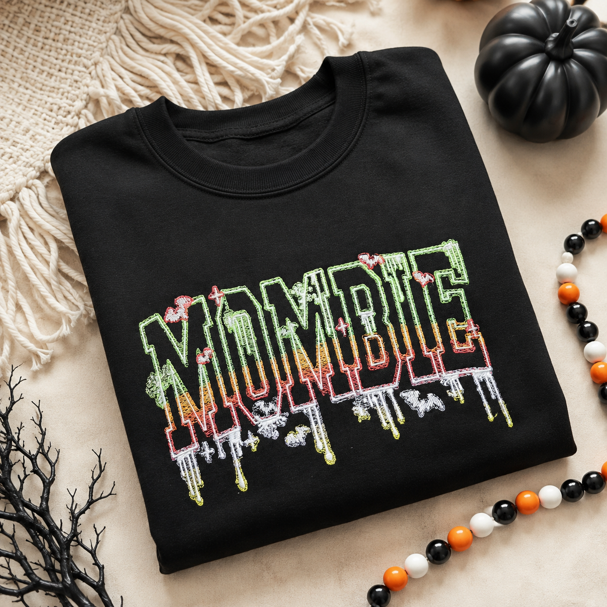 A folded black crewneck t-shirt with a colorful 'MORBIT' graphic in a dripping neon style, styled on a beige fabric background with Halloween props including a black pumpkin, a beaded necklace, and a black branch.