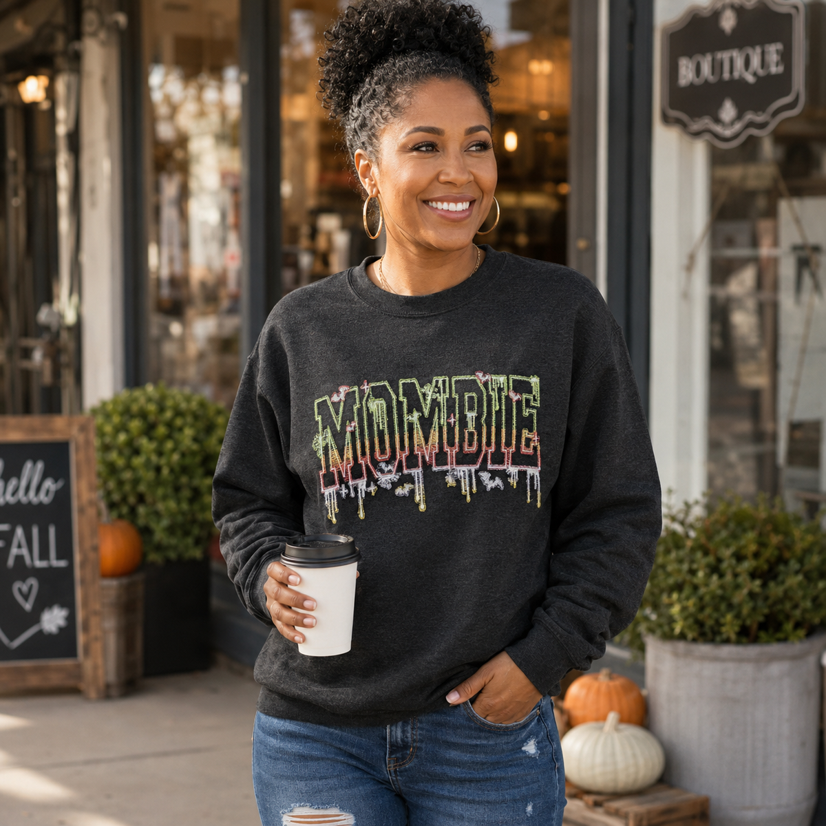 A woman wearing a dark charcoal crewneck sweatshirt with a colorful 'MOMBIE' graphic printed on the front, holding a white coffee cup in front of a boutique storefront.