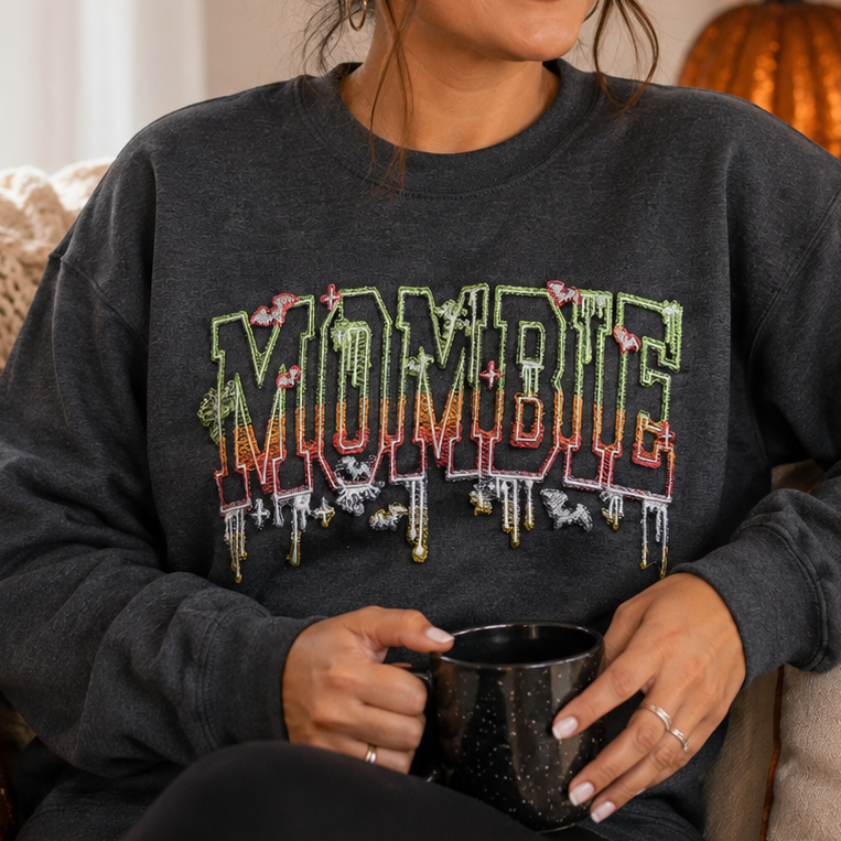 A woman sitting on a couch wearing a dark gray crewneck sweatshirt with a colorful 'MOMBIE' graphic across the chest, holding a black mug in a cozy, decorated living room.