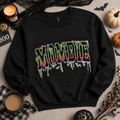 A black crewneck sweatshirt with a colorful 'MOMBITE' graphic in a dripping horror font, styled in a flat lay with Halloween props including pumpkins, a candle, a mug of coffee, and black lace.