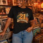 A woman wearing a black graphic t-shirt that reads '21 September Celebrate' in a retro 1970s style, standing in a warm, record-store-like environment with vinyl records and a disco ball in the background.