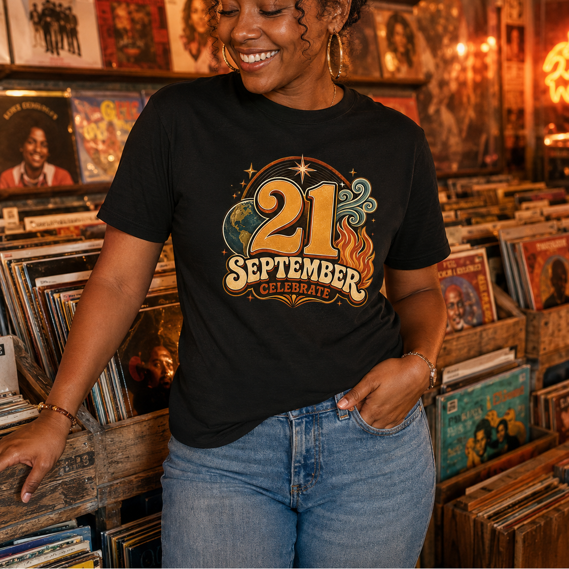 A woman wearing a black graphic t-shirt that reads '21 September Celebrate' in a retro 1970s style, standing in a warm, record-store-like environment with vinyl records and a disco ball in the background.