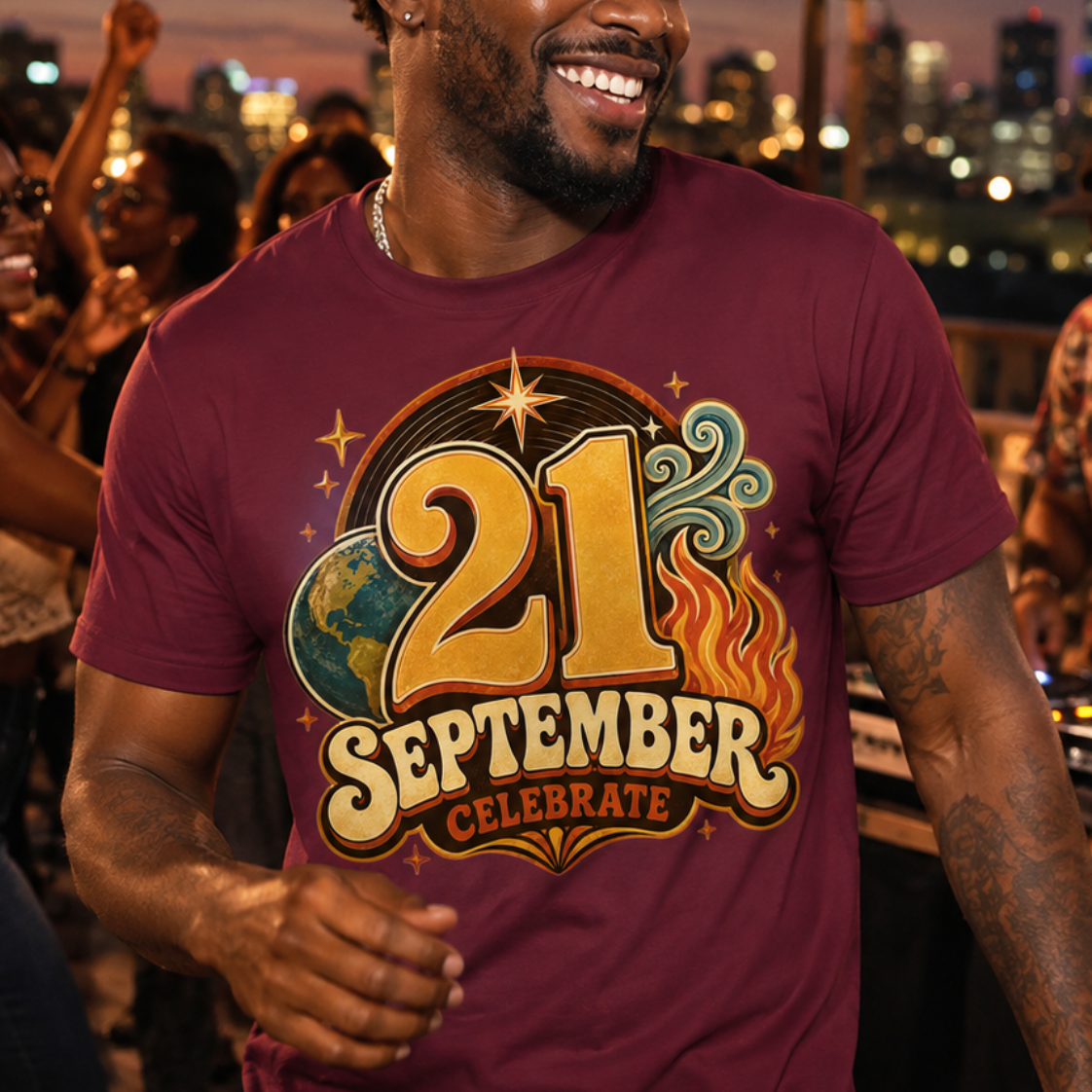 A man smiling on a rooftop party wearing a maroon t-shirt with a '21 September Celebrate' graphic print, with a city skyline and string lights in the background.