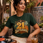 A woman wearing a dark green graphic t-shirt with a retro '21 September Celebrate' design, sitting in a cozy, tiled patio environment with a record player and a drink in the foreground.