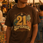 A man wearing a brown graphic t-shirt with a retro '21 September Celebrate' design, standing in a warm, crowded outdoor street market at sunset.