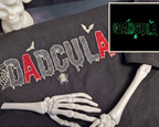 A black graphic sweatshirt featuring the word 'DADCU LA' in a dripping red and gray font with spiderweb and spider motifs. The sweatshirt is styled with skeleton hands and a white skull bust on a dark background.