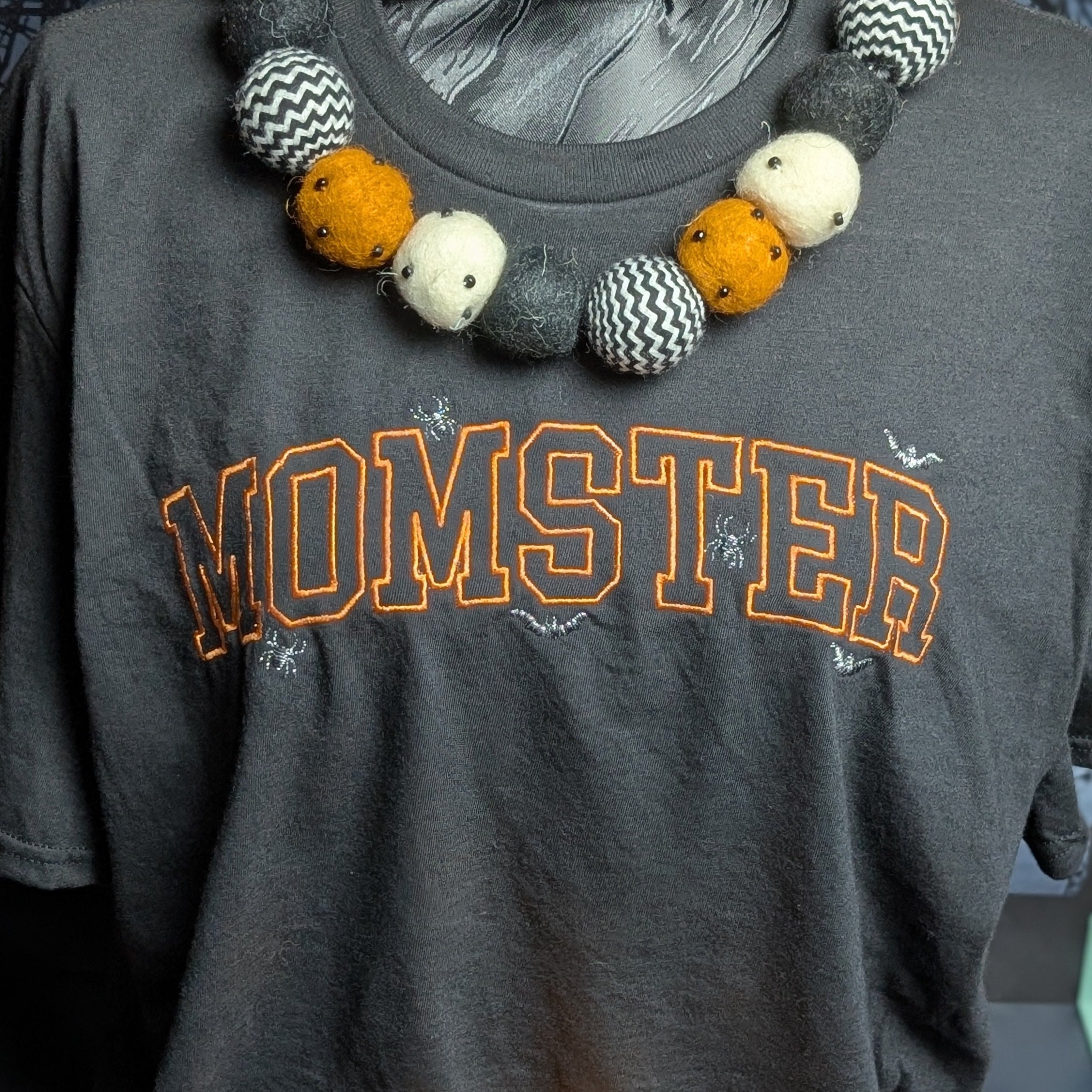 A dark gray graphic t-shirt with 'MOMSTER' printed in orange varsity letters, displayed on a mannequin torso. The shirt is styled with a chunky beaded necklace featuring orange, white, and black-and-white patterned beads, and a matching beaded headband is draped around the mannequin's neck and lower torso.