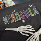 A black short-sleeve T-shirt laid flat with a colorful embroidered 'DAD CULA' design across the chest, styled with skeleton hands and Halloween props.