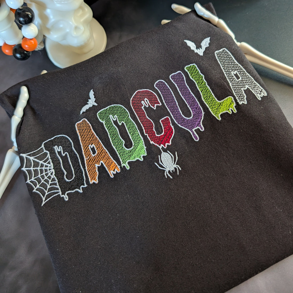 A dark brown drawstring bag with 'DADCOULA' embroidered in colorful, spooky letters with spider and bat motifs, styled next to a white skull decoration.
