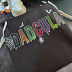 A dark brown drawstring bag with 'DADCOULA' embroidered in colorful, spooky letters with spider and bat motifs, styled next to a white skull decoration.