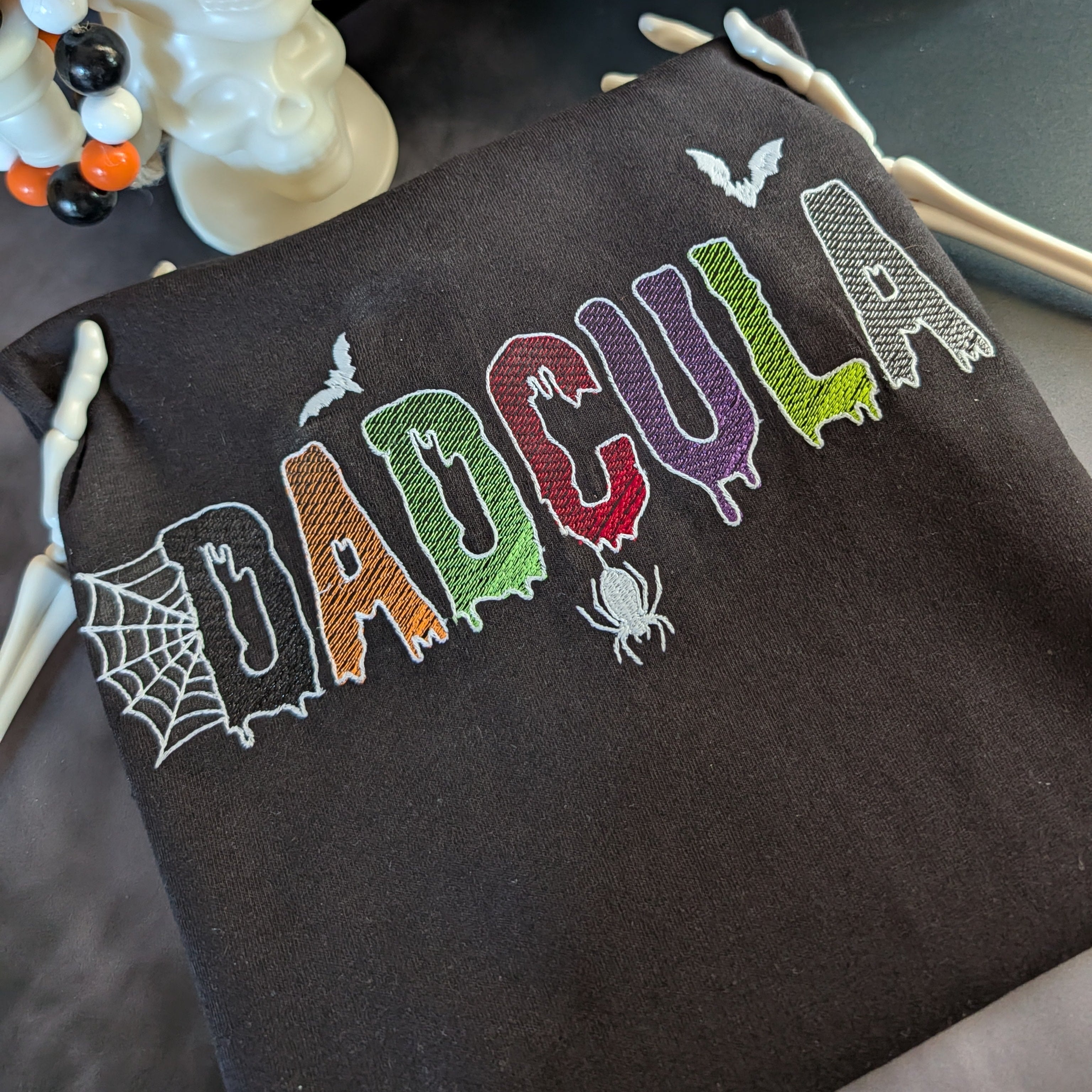 A dark brown drawstring bag with 'DADCOULA' embroidered in colorful, spooky letters with spider and bat motifs, styled next to a white skull decoration.