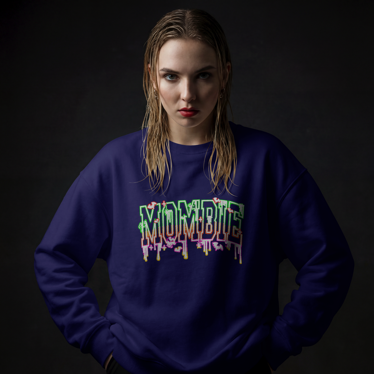 A woman with wet, styled blonde hair wearing a dark navy blue sweatshirt with a colorful dripping 'MOMBIE' graphic on the chest, posed against a dark studio background.